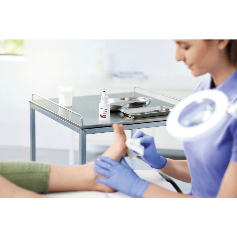 Dans une clinique, une personne portant des gants bleus nettoie le pied d'une autre personne. L'accent est mis ici sur la prévention des infections. Sur la table se trouvent un flacon de 50 ml de spray désinfectant pour plaies Schülke octenisept® de la société Schülke & Mayr GmbH et un plateau métallique contenant des instruments.