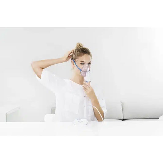 Eine Person benutzt den Auslaufartikel: Beurer kleiner Inhalator IH 60 von Beurer GmbH mit einer Gesichtsmaske an einem weißen Tisch und richtet ihr Haar. Der minimale weiße Hintergrund unterstreicht dieses kompakte Gerät für Atemwegserkrankungen.