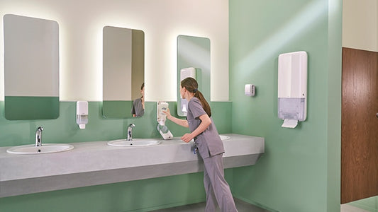 Eine Person in grauem Kittel reinigt ein Waschbecken in einer öffentlichen Toilette mit grünen Wänden, drei Spiegeln und einer Arbeitsplatte mit drei Waschbecken. An der Wand sind Seifen- und Papierhandtuchspender angebracht.