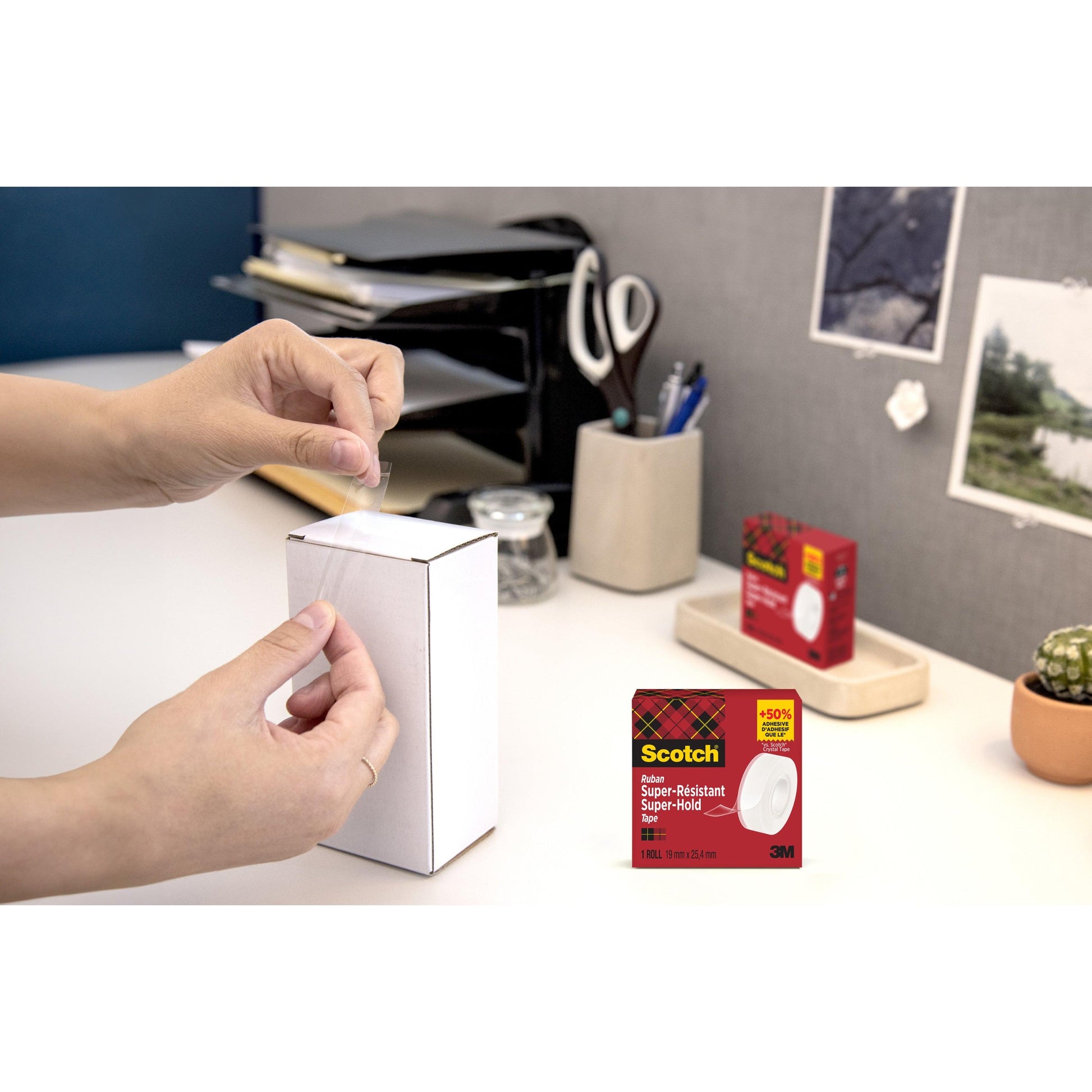 An einem Schreibtisch verschließt jemand eine kleine weiße Schachtel mit Scotch® Super-Hold Secure Tape, 19 mm x 25,4 m (1 Rolle) von 3M Deutschland GmbH. Im Hintergrund sind Büromaterialien, ein Kaktus, Fotos und die Verpackung des Klebebandes zu sehen.
