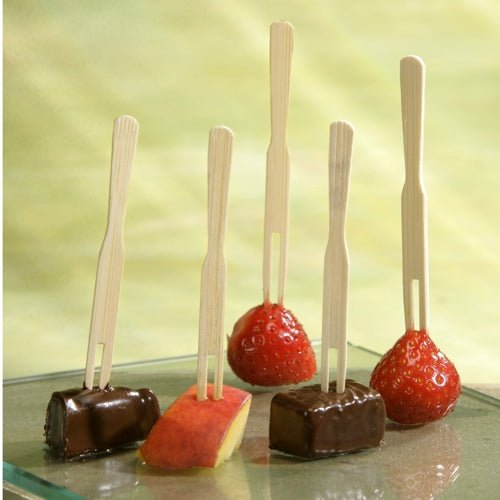 Auf fünf PAPSTAR Fingerfood-Spieße "pure" (8,5 cm) der PAPSTAR GmbH werden zwei Erdbeeren, zwei Schokowürfel und eine Apfelscheibe aufgespießt, die hochkant auf einem Glas vor hellgrünem Hintergrund präsentiert werden.
