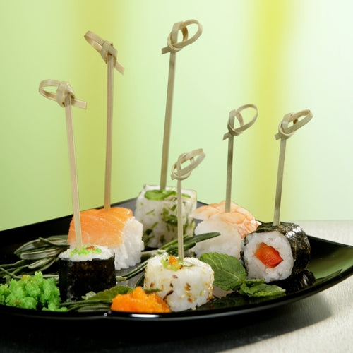 Ein schwarzer Teller mit verschiedenen Sushi, jedes Stück aufgespießt mit PAPSTAR 40 Fingerfood - Picker „pure“ der PAPSTAR GmbH und garniert mit Kräutern und Fischrogen, wird elegant vor einem Hintergrund mit grünem Farbverlauf präsentiert.