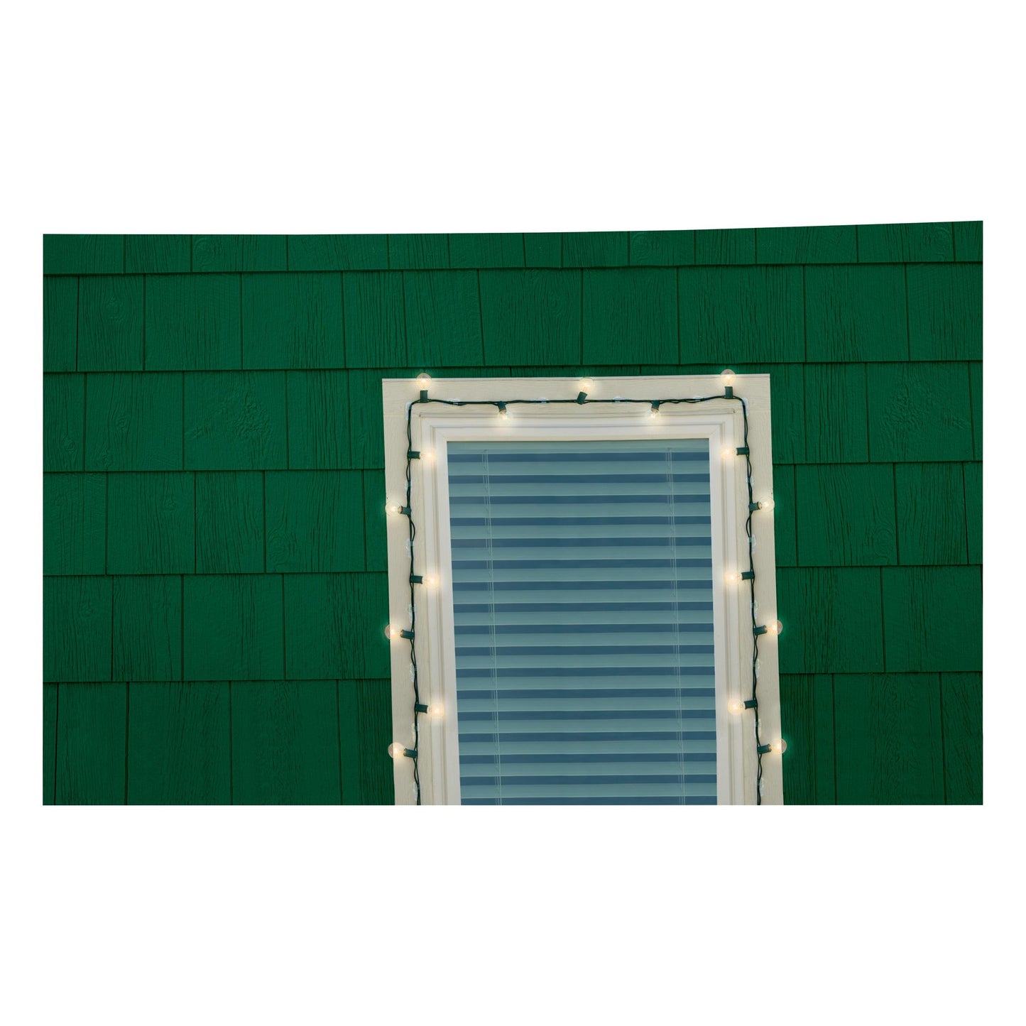 Ein Fenster mit geschlossenen Jalousien an einer grün geschindelten Wand ist mit weißen Lichtern umrandet, die mit 3M Command™ Outdoor Lichter-Clips (17017CLR-AWPU) von 3M Deutschland GmbH für eine zuverlässige Installation der Außenbeleuchtung sicher befestigt wurden.