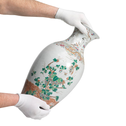 Eine Person mit KleenGuard® G35 Nylon-Handschuhen (24 cm, beidhändig tragbar) von Kimberly-Clark GmbH hält eine große Porzellanvase mit bunten Blumen- und Vogelmotiven vor einem schlichten weißen Hintergrund.