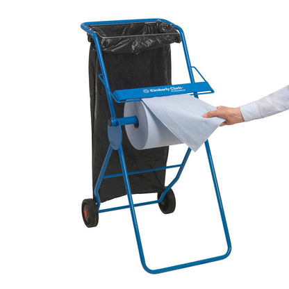 A blue metal stand with two wheels holds a black trash bag and a WypAll® L10 EXTRA wiping cloths large roll (1 roll x 1500 sheets); a person pulls a sheet from the roll. The inscription "Kimberly-Clark GmbH" is affixed on the horizontal surface of the stand.