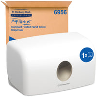 Un distributore bianco Aquarius™ per asciugamani piegati della Kimberly-Clark GmbH (MultiFold, piccolo) è posizionato davanti alla sua confezione marrone. Il contenitore compatto garantisce un prelievo igienico della carta grazie all’apertura rotonda sulla parte anteriore.