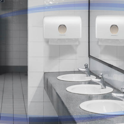 Drei weiße Waschbecken mit silbernen Armaturen sitzen auf einer grauen Arbeitsplatte. Darüber sorgen ein Spiegel und der Kimberly-Clark GmbH Aquarius™ Spender für gefaltete Handtücher (MultiFold/Weiß/Klein) für eine hygienische Handtuchausgabe an der gefliesten Wand.