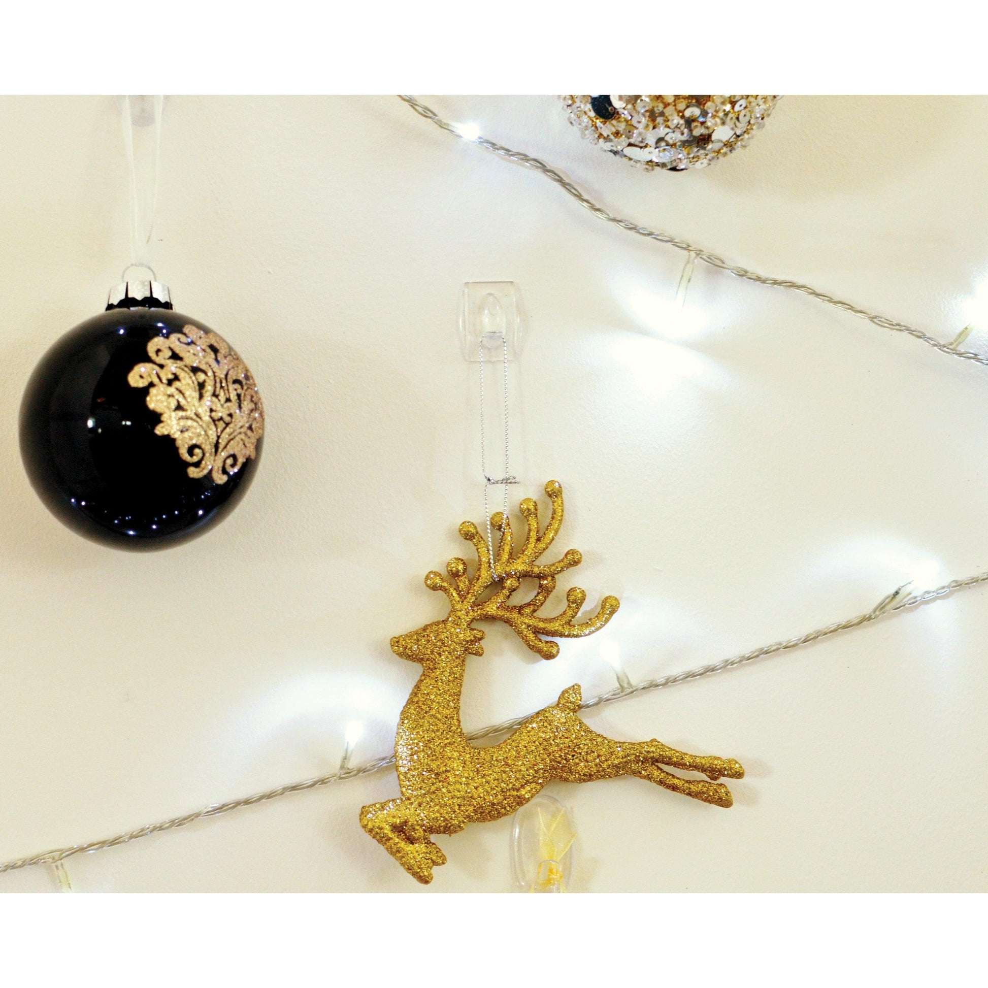 Ein goldglitzerndes Rentierornament wird mit einem transparenten Command™ Mini-Haken der 3M Deutschland GmbH an die Wand gehängt und neben schwarz-goldenen Weihnachtskugeln und weißen Lichterketten präsentiert.