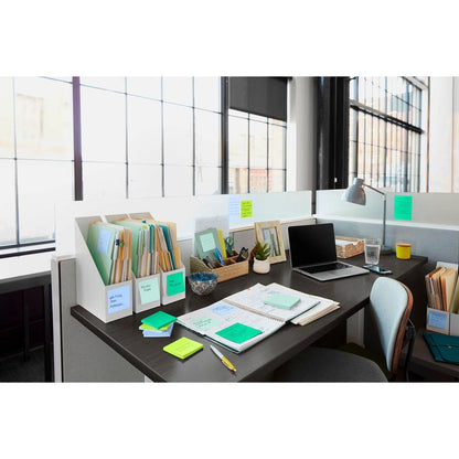 A bright office table with folders, notebooks, stationery, a 3M Deutschland GmbH Post-it® Super Sticky Notes block (76x127mm, 90 sheets, 100% PEFC), a small plant, a lamp, a glass of water, and an open laptop by a large window.