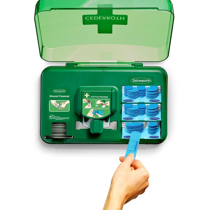 An open wound care dispenser, blue (Orkla Wound Care AB) shows wound cleansing wipes on the left side and blue soft foam dressings on the right side, while a hand uses a blue tool to pull a dressing from the dispenser section.