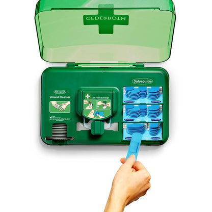 An open wound care dispenser, blue (Orkla Wound Care AB) shows wound cleansing wipes on the left side and blue soft foam dressings on the right side, while a hand uses a blue tool to pull a dressing from the dispenser section.