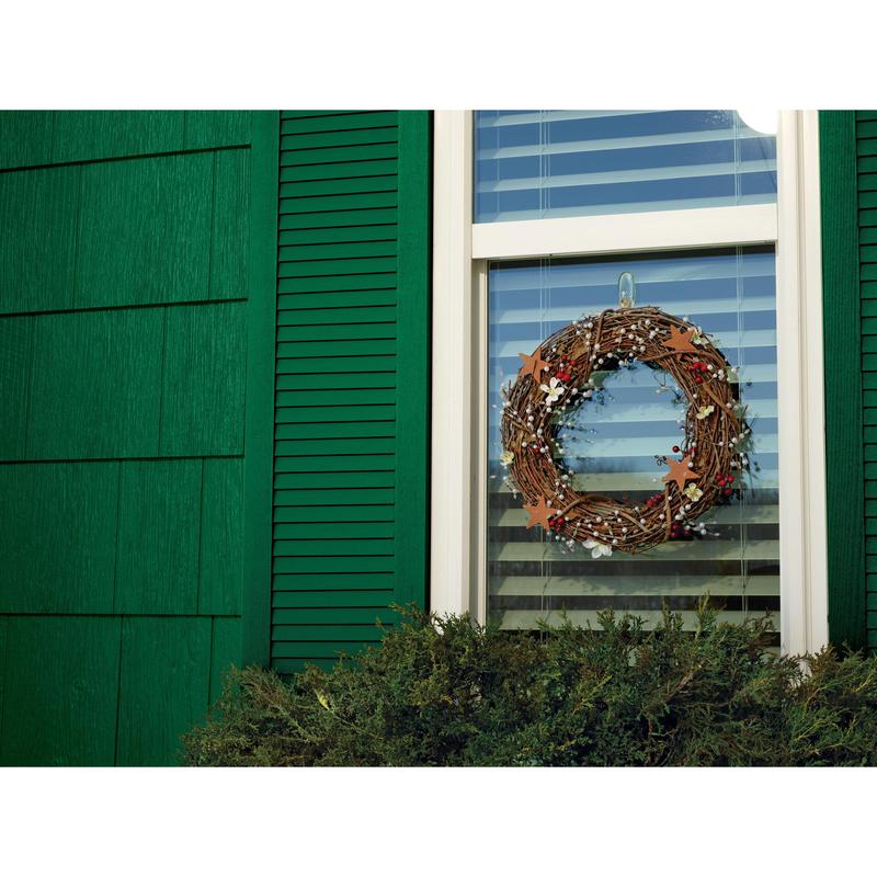 Une couronne de branches avec des feuilles et de petits ornements est suspendue à une fenêtre avec des stores blancs, fixée par un crochet Command™ transparent de taille moyenne pour l'extérieur (ovale, 17091AW) de 3M Deutschland GmbH. Des volets verts encadrent la fenêtre, avec des buissons devant.