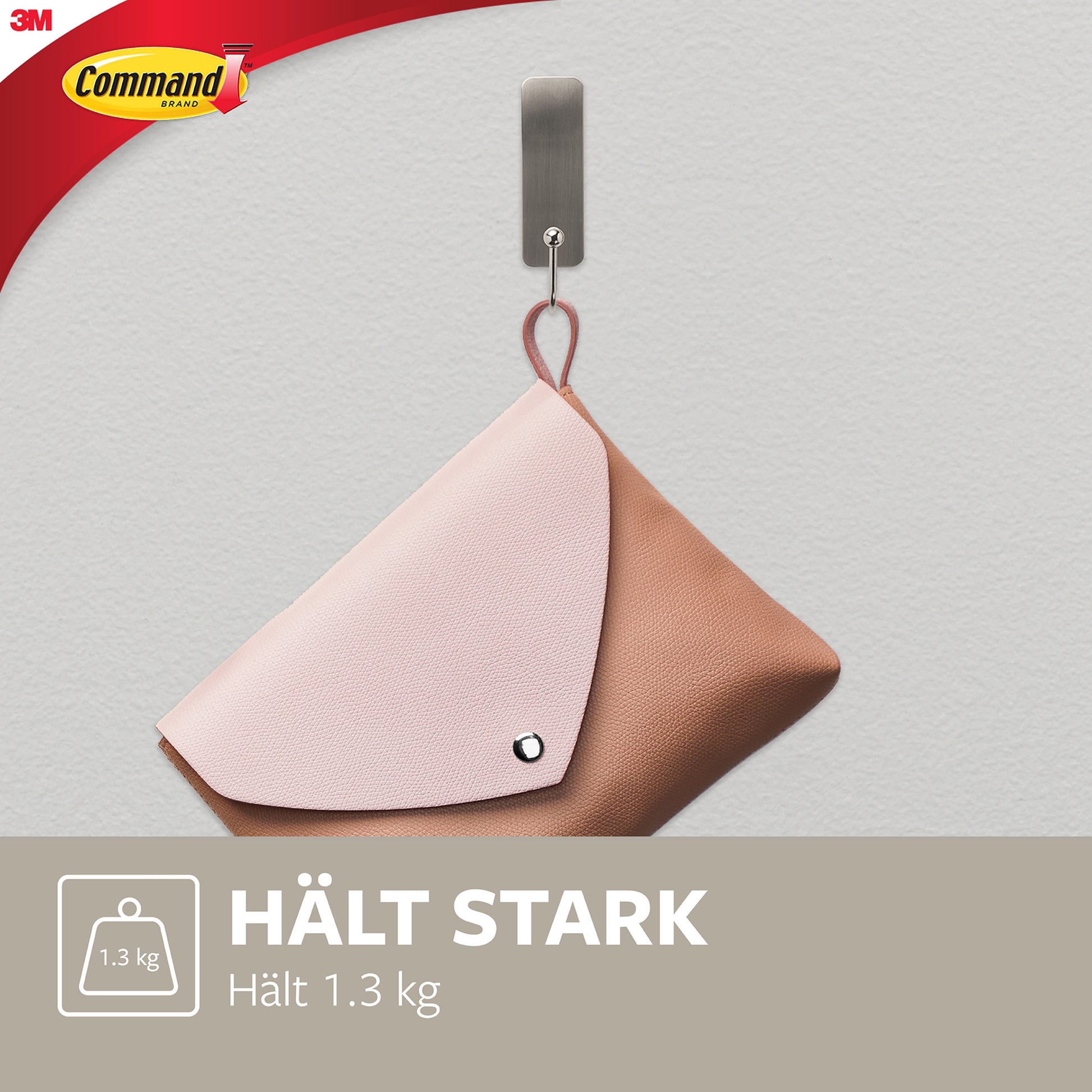 An einem 3M Command™ Mittelgroßer Metallhaken aus gebürstetem Nickel hängt eine braune und rosa Kupplung. Der Text "HÄLT STARK Hält 1,3 kg" mit einem Gewichtssymbol ist abgebildet, und das Logo der 3M Deutschland GmbH erscheint in der oberen linken Ecke.