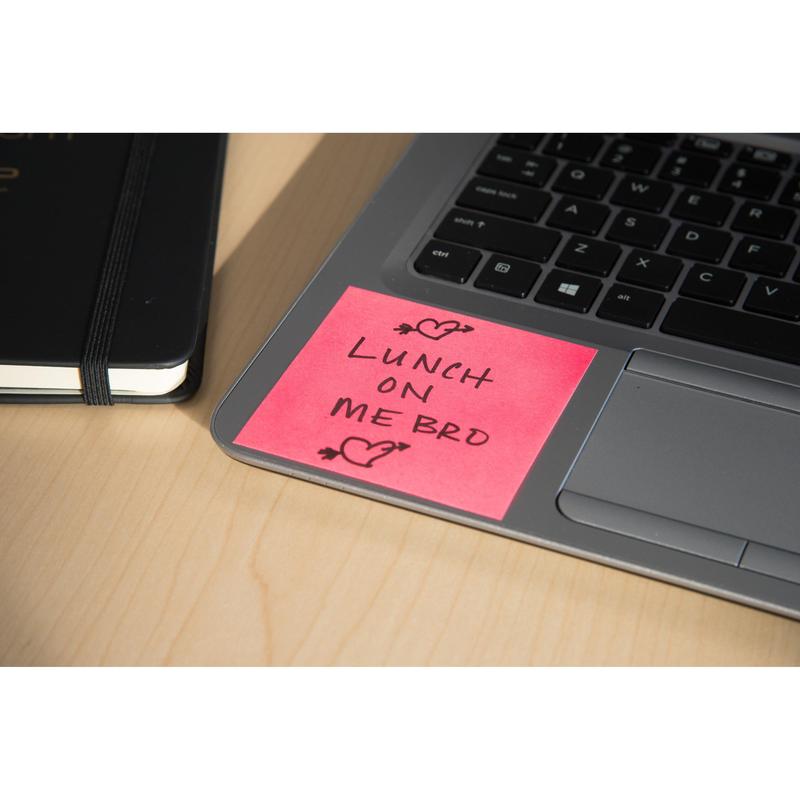 Ein rosafarbener Post-it® Super Sticky Z-Zettel (76x76mm) der 3M Deutschland GmbH mit der Aufschrift "Lunch on me bro" in schwarzer Tinte und zwei Herzen liegt auf einer Laptop-Tastatur neben einem schwarzen Notebook, alles auf einem hellen Holztisch.