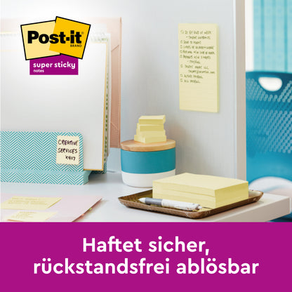 Ein Arbeitsbereich mit gestapelten Post-it® Super Sticky Notes im Großformat, liniert (101 x 101 mm, 45 Blatt/Block, 100% PEFC) von 3M Deutschland GmbH. Ein gelber Zettel hängt an der Wand, Ordner und Büromaterial sind sichtbar. Der deutsche Text lautet: "Haftet sicher, rückstandsfrei ablösbar.