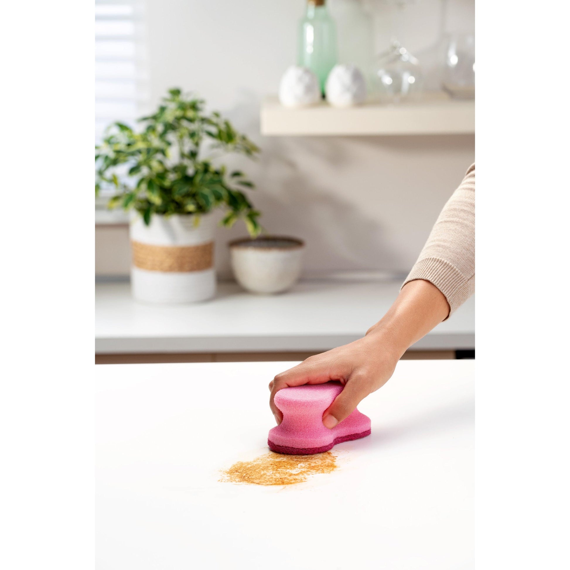 Eine Person benutzt den Scotch-Brite® Delicate Color Comfort nicht kratzender Reinigungsschwamm von 3M Deutschland GmbH, um einen pulverförmigen Fleck auf einer weißen Küchentheke zu reinigen. Im Hintergrund sind Topfpflanzen und Küchenutensilien zu sehen.