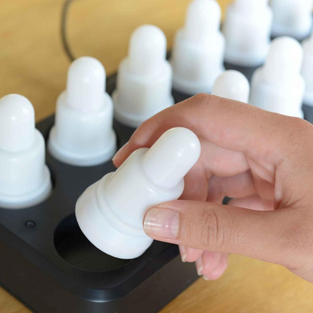 Eine Hand setzt oder entfernt eine weiße, runde LED-Kerze aus dem schwarzen Tablett des Duni GmbH LED Set, Warmweiß & Multicolor - 12 Stück Farbwechsel Leuchten; jede identische, wiederaufladbare Kerze sitzt auf einer Holzunterlage.