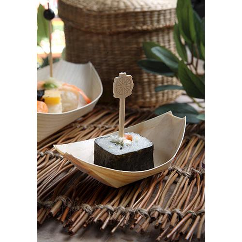 A piece of sushi, wrapped in seaweed and held with a wooden skewer, is served on a PAPSTAR 50 finger food tray wooden "pure" boat from PAPSTAR GmbH, presented on a woven mat with leaves and wicker baskets in the background.