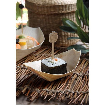 A piece of sushi, wrapped in seaweed and held with a wooden skewer, is served on a PAPSTAR 50 finger food tray wooden "pure" boat from PAPSTAR GmbH, presented on a woven mat with leaves and wicker baskets in the background.