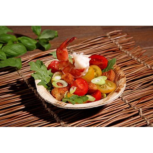 Eine kleine Schüssel mit gekochten Garnelen, Kirschtomaten, Croutons, Rucola und Frühlingszwiebeln wird auf einer gewebten Unterlage in den PAPSTAR 25 Schalen, Palmblatt „pure“ der PAPSTAR GmbH serviert – eine umweltfreundliche Wahl für nachhaltige Veranstaltungen. Basilikumblätter sorgen für einen frischen Touch.
