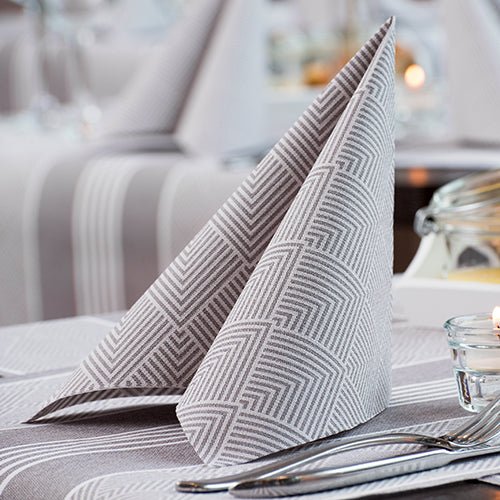 Un tovagliolo piegato con cura PAPSTAR 50 tovaglioli "ROYAL Collection" 1/4 piega 40 x 40 cm grigio della PAPSTAR GmbH sta in piedi su un tavolo con forchetta, coltello, tovaglia a righe, candela e bicchieri sfocati sullo sfondo.
