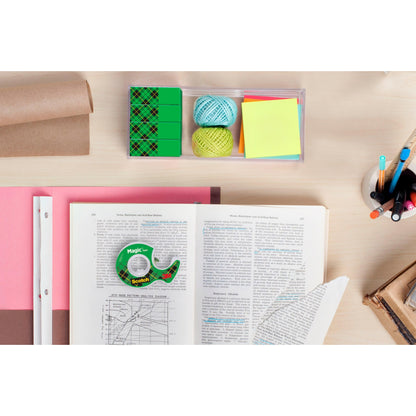 Ein Schreibtisch mit einem aufgeschlagenen Buch, Scotch® Magic™ Unsichtbares Klebeband Vorteilspack (24 Rollen, 3M Deutschland GmbH), farbiges Papier, Haftnotizen, Schnur, Radiergummi, eine Tasse mit Stiften und Bleistiften sowie gestapelte Bücher und gerolltes braunes Papier.