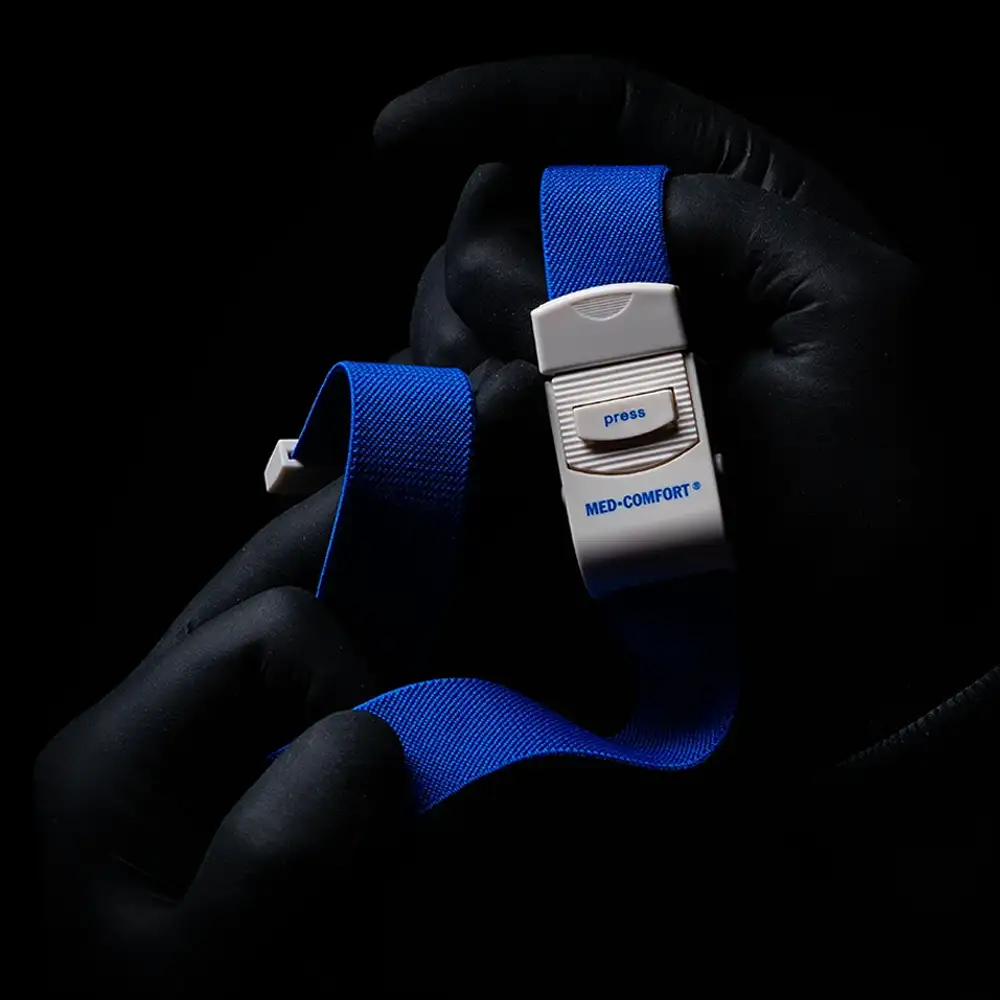 Une personne avec des gants noirs tient un garrot bleu AMPri MED-COMFORT, un instrument indispensable pour les applications médicales. Sur un fond sombre, une boucle en plastique blanc avec un bouton-poussoir est visible.