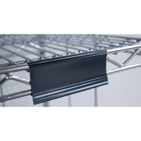 Close-up of a Novocal label frame with paper tags (approx. 100 x 44 mm, 1 piece) attached to a metal wire shelf, with the black plastic label holder on the front edge; the background is out of focus.