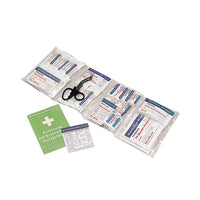 Inside is an open first-aid kit containing various bandaging supplies, medical scissors, a green instruction manual, and a small white guide from the standard contents of the DIN 13157 first-aid kit by Holthaus Medical GmbH & Co. KG, in accordance with the DIN 13157 standard.