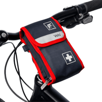 Une petite trousse de premiers secours noire et rouge portant la mention « Holthaus Velo® Trousse de premiers secours pour vélo - 21 pièces | Paquet (1 pièce) » de la société Holthaus Medical GmbH & Co. KG est fixée au guidon d'un vélo. La trousse de premiers secours du vélo est ornée d’une croix blanche et d’un symbole représentant un vélo. On aperçoit également le guidon et une partie du cadre du vélo.