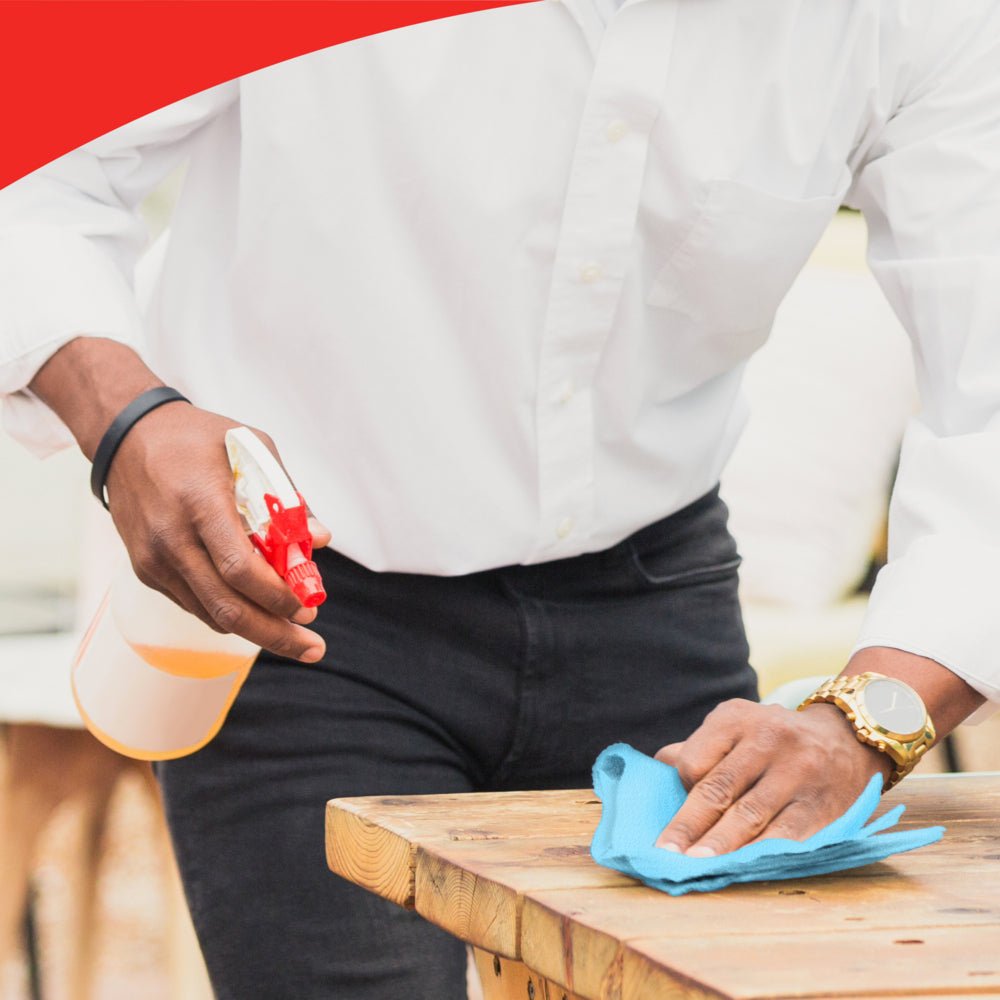 Una persona con camicia bianca e orologio dorato pulisce un tavolo di legno con un rotolo blu WypAll® X60 General Clean di Kimberly-Clark GmbH. Il suo volto non è visibile. L'immagine ha un bordo rosso nella parte superiore.