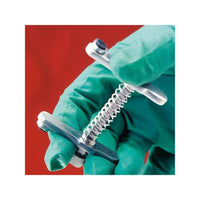 A person wearing green gloves is holding, against a red background, an Altruan Servoprax muzzle for cats with a cylindrical handle and a threaded screw, which is typically used by veterinarians for veterinary procedures.