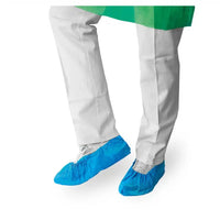 Une personne vêtue d'une blouse de laboratoire blanche et de couvre-chaussures bleus jetables de la marque Meditrade GmbH se tient devant un fond blanc ; seules ses jambes sont visibles.