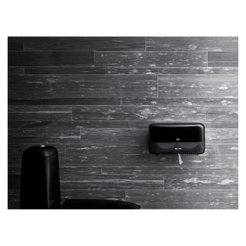 Ein modernes Badezimmer mit dunkelgrauen Holzdielenwänden, einem an der Wand befestigten schwarzen Toilettenpapierspender und dem Rand einer schwarzen Toilette in der unteren linken Ecke. Aus dem Spender ragt an der Unterseite ein Stück Tork 472199 hülsenloses Midi Toilettenpapier Advanced Weiß T7 2-lagig | Karton (36 Rollen) von TORK heraus.