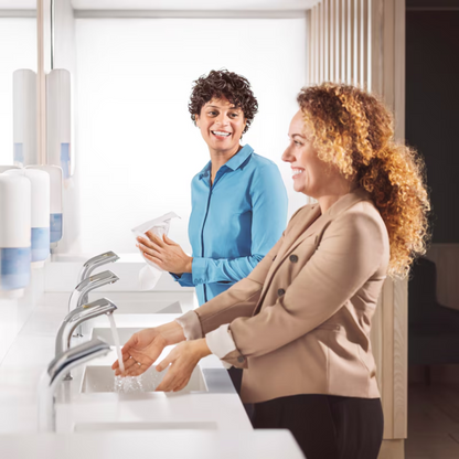 Two women are standing at the sinks of a public restroom. One is washing her hands with Tork 520201 Pure Hand Foam Soap Premium S4 (1000 ml) from TORK – Essity Professional Hygiene Germany GmbH, the other is smiling and holding paper towels in her hand. Soap dispensers are on the washstand.