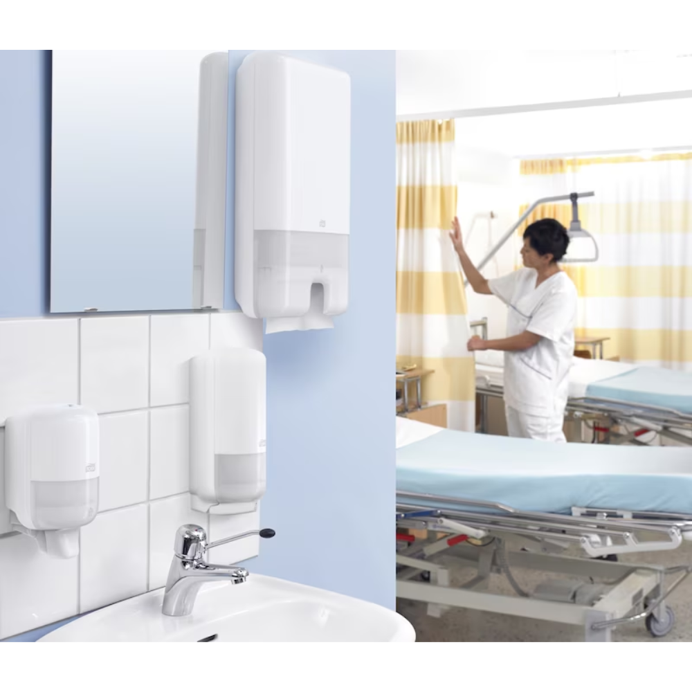 In einem Krankenhauszimmer steht ein Arzt in weißer Uniform neben einem Bett. Im Vordergrund ist ein Waschbecken mit einem Tork Xpress® 552000 Spender für Multifold Handtücher H2 von TORK – Essity Professional Hygiene Germany GmbH ausgestattet. Das Zimmer ist mit medizinischen Geräten und gelb gestreiften Vorhängen ausgestattet.