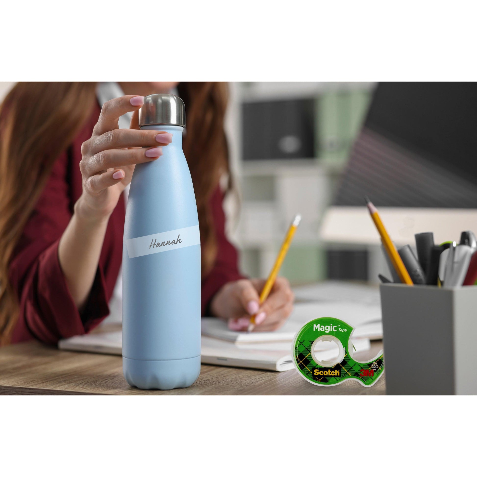 Eine Person an einem Schreibtisch hält eine hellblaue Wasserflasche mit der Aufschrift "Hannah". Im Vordergrund sind eine Scotch® Magic™ Unsichtbares Klebeband Nachfüllpackung (2 Rollen, 19 mm x 7,5 m) der 3M Deutschland GmbH und Büromaterial zu sehen.