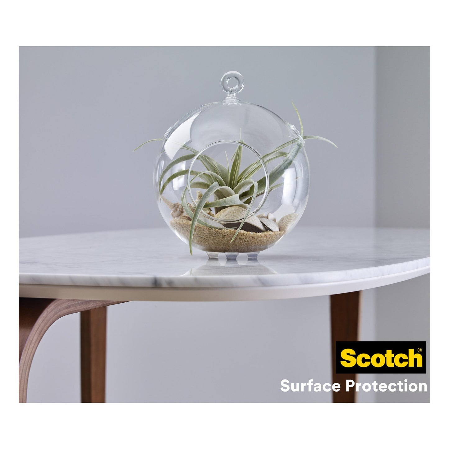 Ein Glasterrarium mit Luftpflanzen und Sand steht auf einer weißen Marmortischplatte mit Holzbeinen. In der Ecke des Tisches steht das Logo der 3M Deutschland GmbH und "Scotch® Stoßabsorbierende Elastikpuffer, selbsthaftend, transparent".