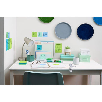 A tidy white desk with a lamp, folders, notebooks, pens, cups, and glasses. The Post-it® Super Sticky Notes (76 mm x 127 mm) from 3M Deutschland GmbH hang on the wall with colorful plates. A chair stands in front of the desk.