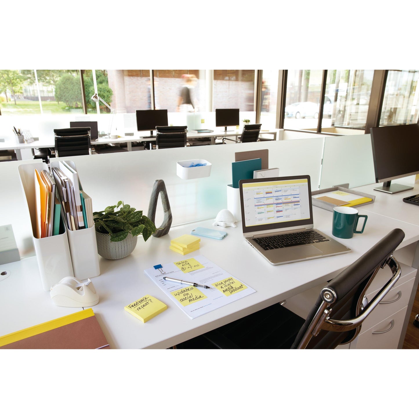 A modern open-plan office with a desk featuring a laptop, 3M Post-it® Notes Yellow (76x102 mm, 100 sheets/block), documents, a coffee mug, supplies, and a plant. Large windows fill the space with light; outside, a blurred figure passes by.