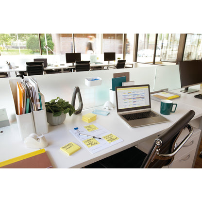 A modern open-plan office with a desk featuring a laptop, 3M Post-it® Notes Yellow (76x102 mm, 100 sheets/block), documents, a coffee mug, supplies, and a plant. Large windows fill the space with light; outside, a blurred figure passes by.