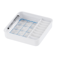 A square organizer made of white plastic with multiple compartments, ideal for storing medications, featuring a mesh section for small items and a pen holder on the side. The mesh section is designed for the Melipul cup-shaped tablet from Helmut Schwarz GmbH, complete with small, round, transparent lids for neat storage.